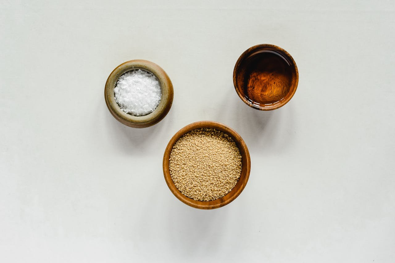 Top view of sesame seeds, salt, and oil in wooden bowls on a white background, ideal for culinary themes.